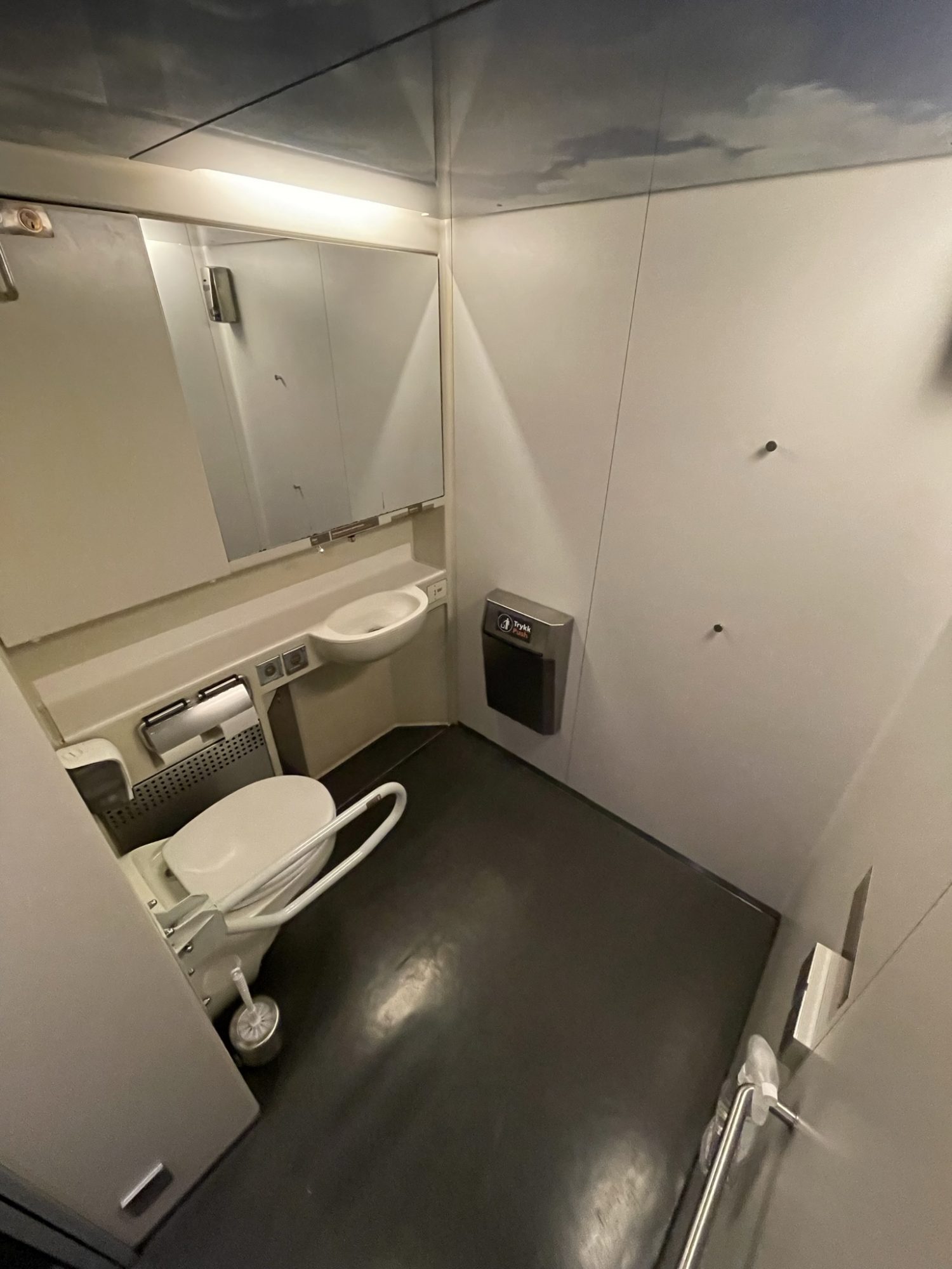 Et kvadratisk toalettrom. Vegghengt toalett til venstre med vippbart armlene på høyre side.